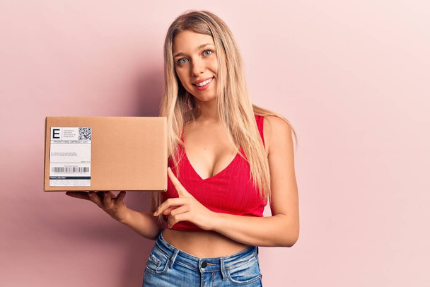 Young beautiful blonde woman holding delivery package looking positive and happy standing and smiling with a confident smile showing teeth  - Foto, afbeelding