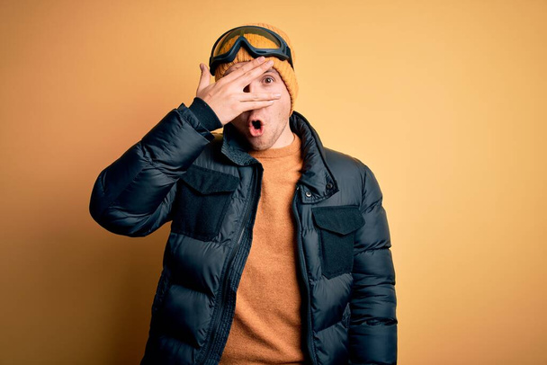 Young handsome caucasian man wearing hat, coat and ski glasses for winter and snow weather peeking in shock covering face and eyes with hand, looking through fingers with embarrassed expression. - Foto, Imagen