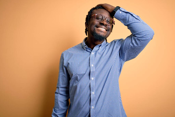 Young handsome african american man wearing shirt and glasses over yellow background smiling confident touching hair with hand up gesture, posing attractive and fashionable - Fotó, kép