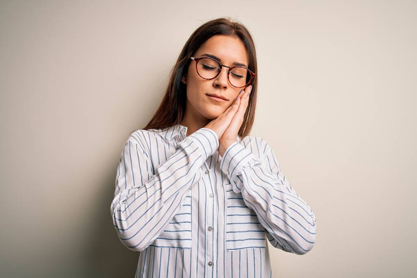Young beautiful brunette woman wearing casual shirt and glasses over white background sleeping tired dreaming and posing with hands together while smiling with closed eyes. - Foto, Imagen