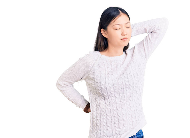 Νεαρή όμορφη Κινέζα γυναίκα φορώντας casual πουλόβερ που υποφέρει από τραυματισμό του αυχένα, αγγίζοντας το λαιμό με το χέρι, μυϊκός πόνος  - Φωτογραφία, εικόνα