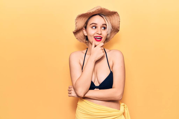 Young beautiful blonde woman wearing bikini and summer hat with hand on chin thinking about question, pensive expression. smiling with thoughtful face. doubt concept.  - Fotoğraf, Görsel