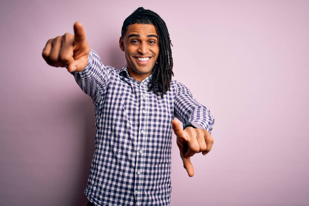 Junger gut aussehender afro-amerikanischer Afro-Mann mit Dreadlocks, der lässig Hemd trägt und mit den Fingern in die Kamera zeigt, positiv und fröhlich lächelt - Foto, Bild