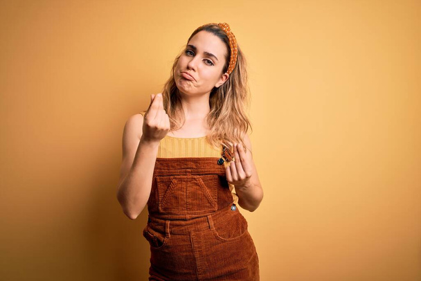 Young beautiful blonde woman wearing overalls and diadem standing over yellow background doing money gesture with hands, asking for salary payment, millionaire business - Photo, Image