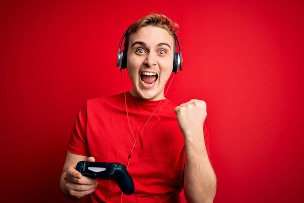 Giovane bel giocatore rossa uomo che gioca al videogioco con cuffie e joystick urlando orgoglioso, celebrando la vittoria e il successo molto eccitato con le braccia alzate - Foto, immagini