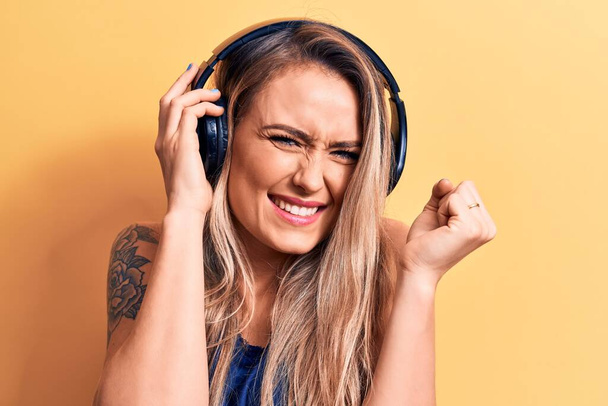 Young beautiful blonde woman listening to music using headphones over yellow background screaming proud, celebrating victory and success very excited with raised arm - Zdjęcie, obraz