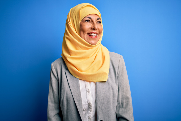 Mulher de negócios morena de meia idade vestindo hijab tradicional muçulmano sobre fundo azul olhando para o lado com sorriso no rosto, expressão natural. Rindo confiante
. - Foto, Imagem