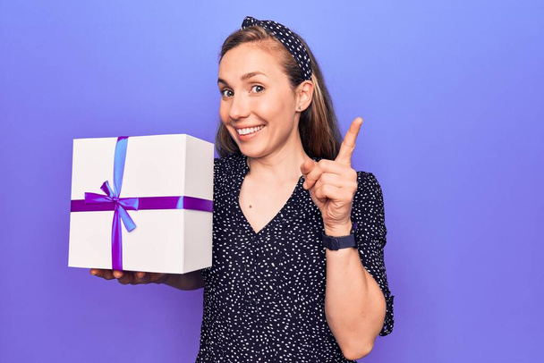 Fiatal gyönyörű szőke nő kezében születésnapi ajándék doboz mosolygós boldog mutató kéz és ujj oldalra  - Fotó, kép