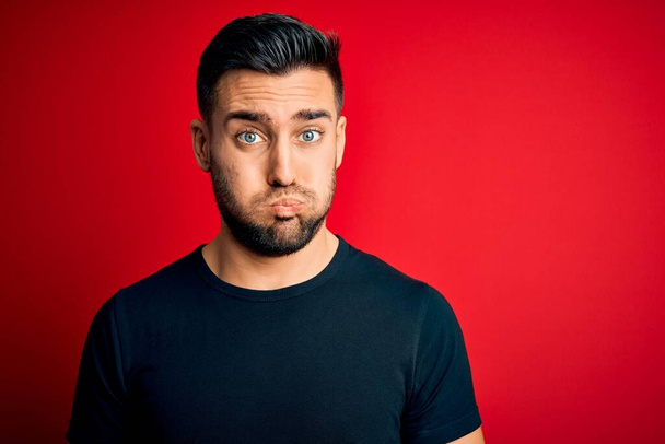 Young handsome man wearing casual black t-shirt standing over isolated red background puffing cheeks with funny face. Mouth inflated with air, crazy expression. - Photo, Image