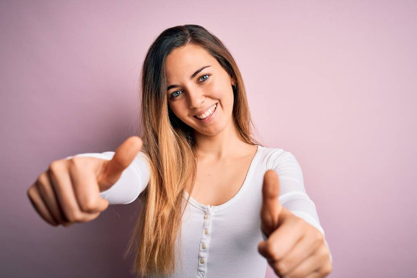 Young beautiful blonde woman with blue eyes wearing white t-shirt over pink background approving doing positive gesture with hand, thumbs up smiling and happy for success. Winner gesture. - Фото, изображение