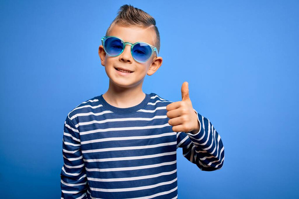 Jeune enfant caucasien aux yeux bleus portant des lunettes de soleil sur fond bleu faisant des pouces heureux geste avec la main. Approuver l'expression en regardant la caméra montrant le succès. - Photo, image
