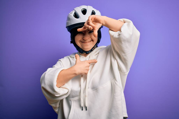 Giovane donna ciclista sindrome di Down indossando casco bici di sicurezza su sfondo viola sorridente facendo telaio con le mani e le dita con il viso felice. Creatività e concetto di fotografia. - Foto, immagini
