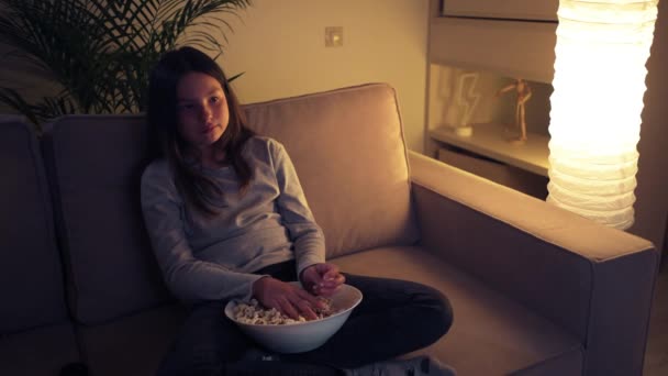 A beautiful girl watches TV in the evening and throws popcorn out of fright. - Video, Çekim