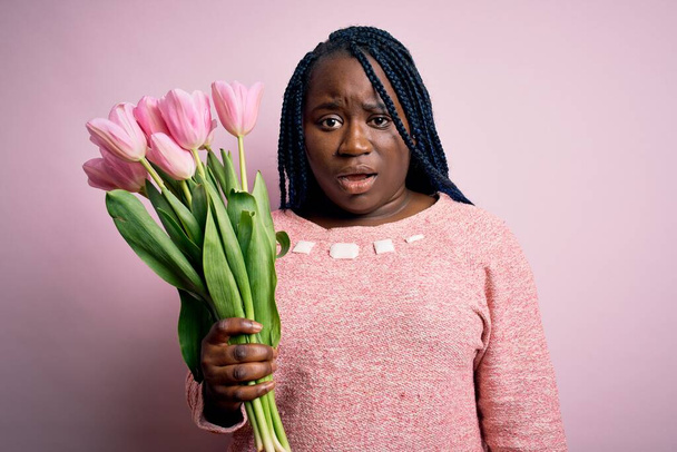 Giovane afroamericana plus size donna con trecce in possesso di bouquet di fiori di tulipani rosa In faccia shock, cercando scettico e sarcastico, sorpreso a bocca aperta - Foto, immagini