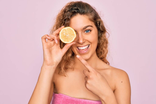 Beautiful woman with blue eyes wearing towel shower after bath holding lemon fruit over eye very happy pointing with hand and finger - Photo, Image