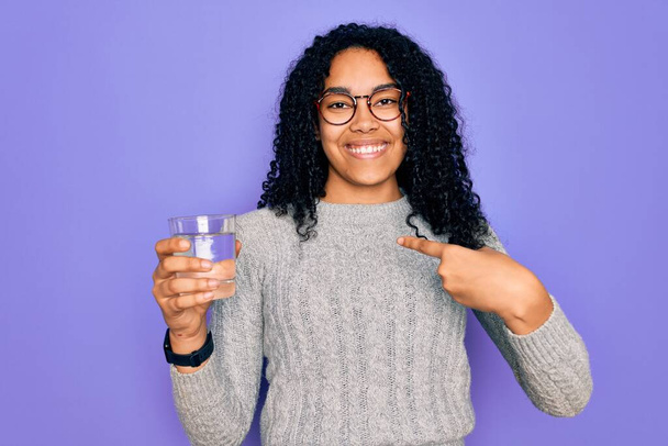 Молодая африканская американка пьет стакан воды на изолированном фиолетовом фоне с удивленным лицом, указывающим пальцем на себя
 - Фото, изображение