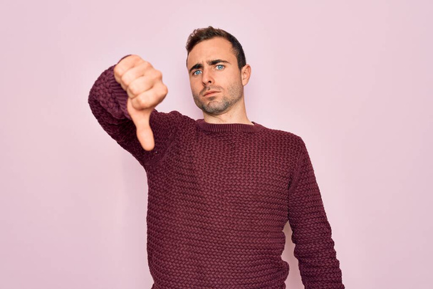 Young handsome man with blue eyes wearing casual sweater standing over pink background looking unhappy and angry showing rejection and negative with thumbs down gesture. Bad expression. - Valokuva, kuva