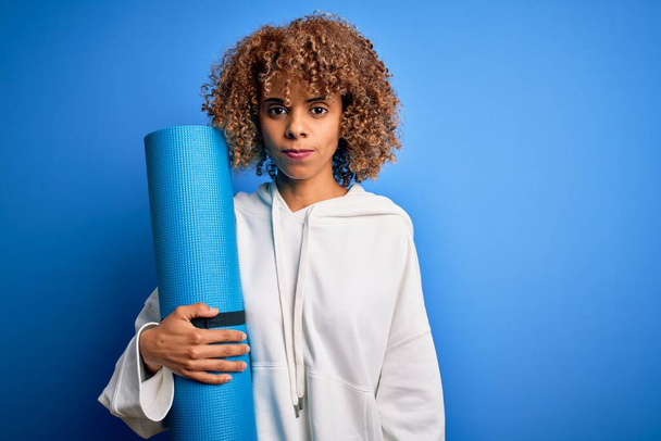 Junge afrikanisch-amerikanische Frau, die Sport treibt, hält Yogamatte über isoliertem blauem Hintergrund mit selbstbewusstem Gesichtsausdruck - Foto, Bild