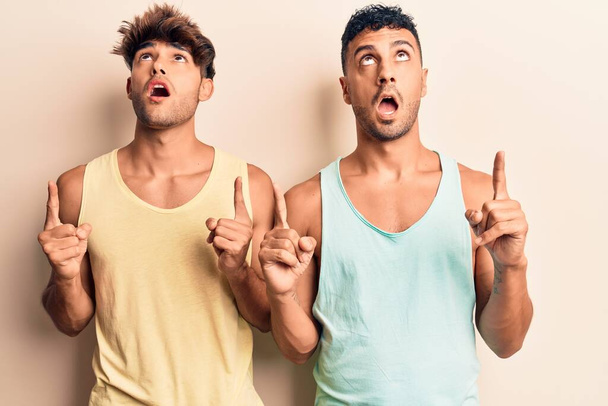 Mladý gayové pár na sobě ležérní oblečení užaslý a překvapený vzhlíží a ukazuje s prsty a zvednutými pažemi.  - Fotografie, Obrázek