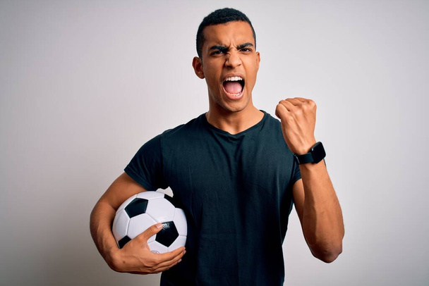 Yakışıklı afro-amerikan adam futbol topunu beyaz arka planda tutarken kızgın ve kızgın yumruk kaldırırken öfkeyle bağırıyor. Öfke ve agresif kavram. - Fotoğraf, Görsel