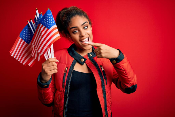 Jovem menina patriótica afro-americana segurando bandeiras dos Estados Unidos sobre fundo vermelho isolado muito feliz apontando com a mão e o dedo
 - Foto, Imagem