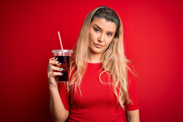 Genç, güzel, sarışın bir kadın kırmızı arka planda saman kullanarak kola içerken akıllı bir yüz ifadesiyle ciddi ciddi düşünüyor. - Fotoğraf, Görsel