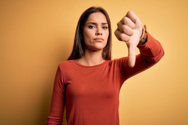 Young beautiful brunette woman wearing casual t-shirt standing over yellow background looking unhappy and angry showing rejection and negative with thumbs down gesture. Bad expression. - Photo, Image