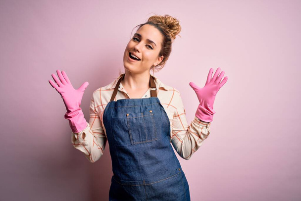 Νεαρή όμορφη ξανθιά καθαρίστρια που κάνει οικιακές εργασίες φορώντας arpon και γάντια γιορτάζει τρελό και έκπληκτος για την επιτυχία με τα χέρια υψωμένα και ανοιχτά μάτια ουρλιάζοντας ενθουσιασμένος. Έννοια νικητή - Φωτογραφία, εικόνα