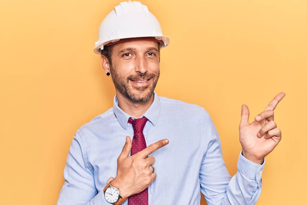 Mladý pohledný muž v architektově klobouku, usmívá se a dívá se na kameru ukazující oběma rukama a prsty do strany.  - Fotografie, Obrázek