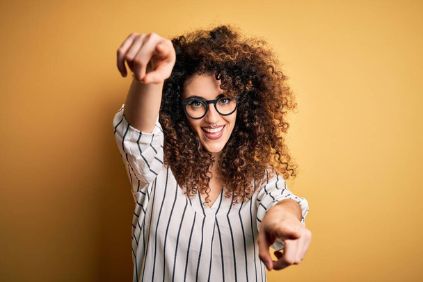Young beautiful woman with curly hair and piercing wearing striped shirt and glasses pointing to you and the camera with fingers, smiling positive and cheerful - Photo, Image