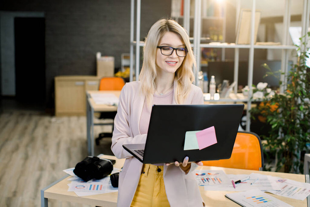 Concentrated young beautiful blond Caucasian businesswoman working on laptop in bright modern office. Pretty woman in glasses working on laptop in the office to finish the project - Photo, image