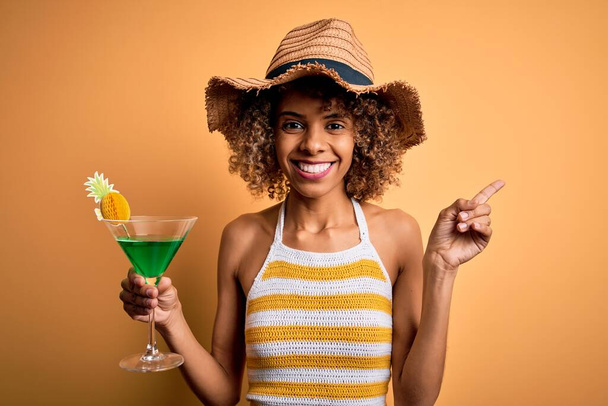 afro-américaine touriste femme avec bouclé en vacances portant chapeau d'été cocktail boire très heureux pointant avec la main et le doigt sur le côté - Photo, image