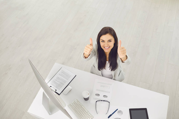 Ansicht von oben. Positiv selbstbewusste Geschäftsfrau lächelt und hebt den Daumen, während sie am Tisch am Arbeitsplatz im Büro steht. - Foto, Bild