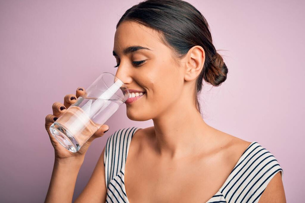 Young beautiful brunette woman smiling happy and confident. Standing with smile on face drinking glass of healthy water to refreshment over isolated pink background - Zdjęcie, obraz