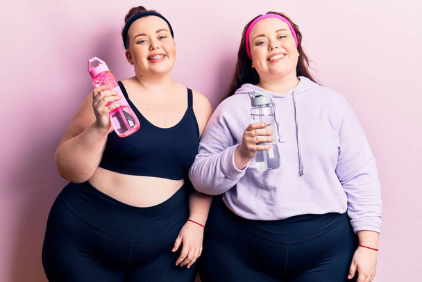 Junge Plus-Size-Zwillinge in Sportbekleidung, die eine Flasche Wasser trinken, sehen im Stehen positiv und glücklich aus und lächeln mit einem selbstbewussten Lächeln, das Zähne zeigt  - Foto, Bild