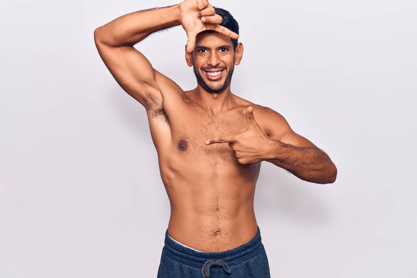 Young latin man standing shirtless smiling making frame with hands and fingers with happy face. creativity and photography concept.  - 写真・画像
