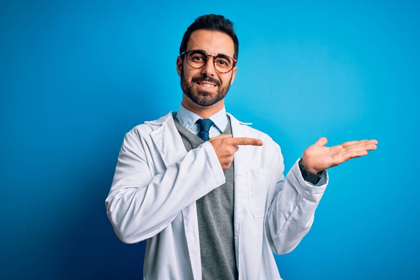 Young handsome doctor man with beard wearing coat and glasses over blue background amazed and smiling to the camera while presenting with hand and pointing with finger. - Photo, Image