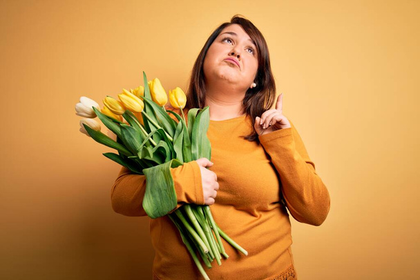 Bella plus size donna in possesso di bouquet romantico di tulipani naturali fiori su sfondo giallo Indicando un aspetto triste e sconvolto, indicando la direzione con le dita, infelice e depresso. - Foto, immagini