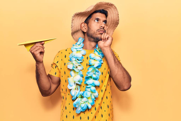 Jovem latino vestindo chapéu de verão e hawaiian lei segurando papel avião rosto sério pensando em questão com a mão no queixo, pensativo sobre ideia confusa
  - Foto, Imagem