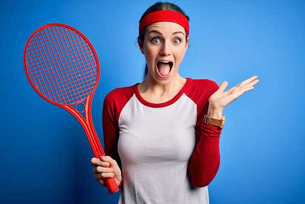 Fiatal gyönyörű vörös hajú sportnő teniszezik ütő kék háttér nagyon boldog és izgatott, győztes kifejezés ünneplő győzelem sikoltozik nagy mosollyal és felemelt kézzel - Fotó, kép