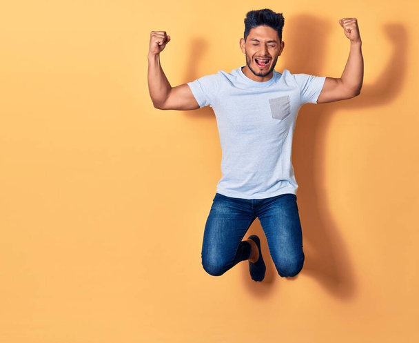 Junger gutaussehender lateinischer Mann in lässiger Kleidung, der glücklich lächelt. Mit einem Lächeln im Gesicht hüpfen und Siegerzeichen mit erhobenen Fäusten vor gelbem Hintergrund machen - Foto, Bild