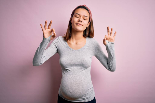 Young beautiful teenager girl pregnant expecting baby over isolated pink background relaxed and smiling with eyes closed doing meditation gesture with fingers. Yoga concept. - Photo, Image