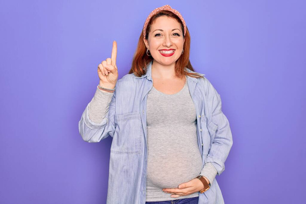 Jovem mulher grávida ruiva bonita esperando bebê sobre fundo roxo isolado mostrando e apontando para cima com o dedo número um, sorrindo confiante e feliz
. - Foto, Imagem