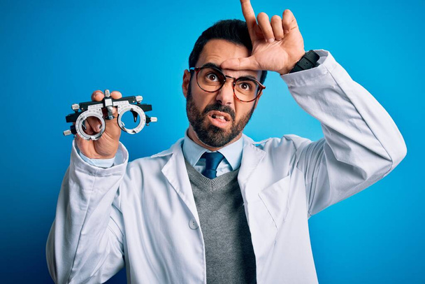 Jeune homme optique beau avec barbe tenant des lunettes d'optométrie sur fond bleu se moquant des gens avec les doigts sur le front faisant geste de perdant se moquant et insultant. - Photo, image