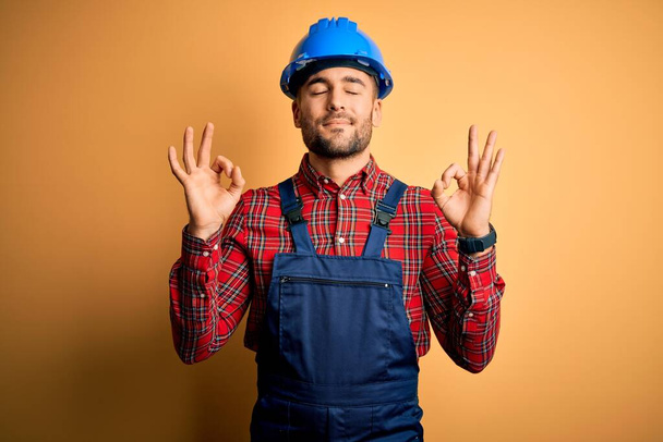 Молодой строитель в строительной форме и защитном шлеме на жёлтом фоне расслабляется и улыбается с закрытыми глазами, делая медитативный жест пальцами. Концепция йоги
. - Фото, изображение