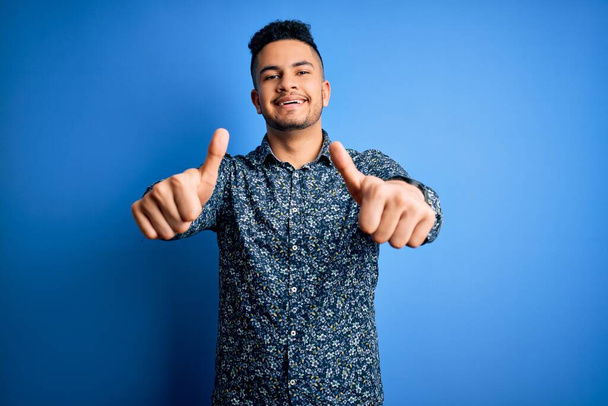 Young handsome man wearing casual shirt standing over isolated blue background approving doing positive gesture with hand, thumbs up smiling and happy for success. Winner gesture. - Photo, Image
