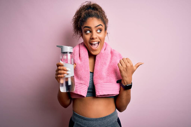 Νεαρό Αφροαμερικανό σπορ κορίτσι που κάνει αθλητισμό φορώντας πετσέτα πίνοντας μπουκάλι νερό δείχνοντας και δείχνοντας με τον αντίχειρα μέχρι το πλάι με χαρούμενο πρόσωπο χαμογελώντας - Φωτογραφία, εικόνα