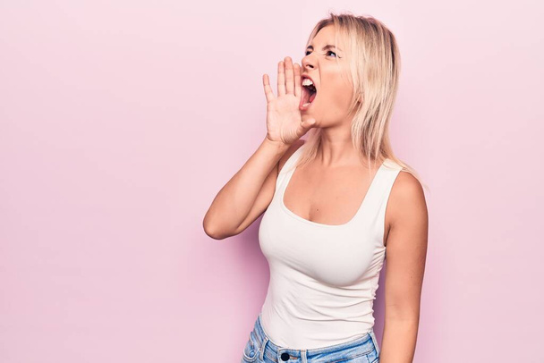 Молода красива блондинка в повсякденній футболці стоїть над ізольованим рожевим фоном і кричить голосно на бік з рукою на рот. Концепція комунікації
. - Фото, зображення