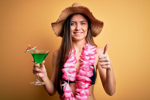Beautiful woman with blue eyes on vacation wearing bikini and hawaiian lei drinking cocktail happy with big smile doing ok sign, thumb up with fingers, excellent sign - Photo, Image