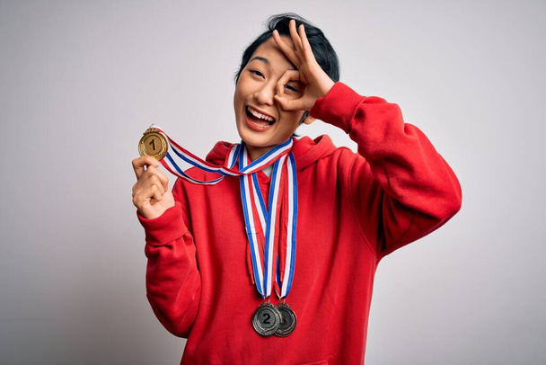 Молодая красивая азиатка победительница носит медали на изолированном белом фоне с счастливым лицом улыбается делает хорошо знак с рукой на глазу глядя сквозь пальцы
 - Фото, изображение
